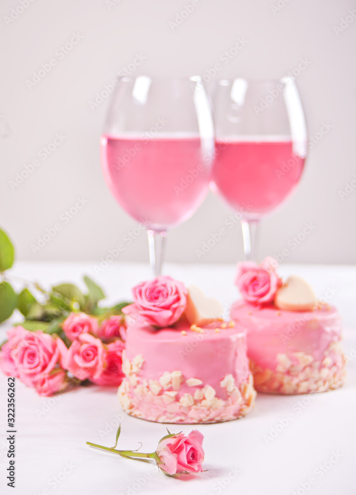 Two mini cakes, glasses with pink grape wine and rose flowers on a white table. Romantic dinner concept Two mini cakes, glasses with pink grape wine and rose flowers on a white table. Romantic dinner concept