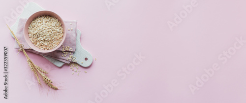Bowl of dry oat flakes with ears of wheat on light pink background