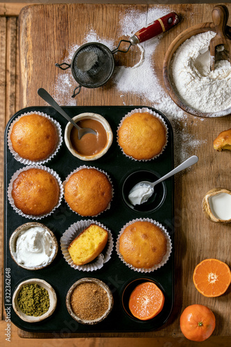 Homemade citrus oranges or clementines sweet muffins cakes in baked tray with flour, ingredients and different topping above over wooden cutting board as background. Flat lay, space