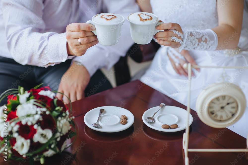 The bride and groom are sitting in a cafe with cups of coffee with foam in the form of a heart and a bouquet of roses and eustomas. Photography, concept. The bride and groom are sitting in a cafe with cups of coffee with foam in the form of a heart and a bouquet of roses and eustomas. Photography, concept.