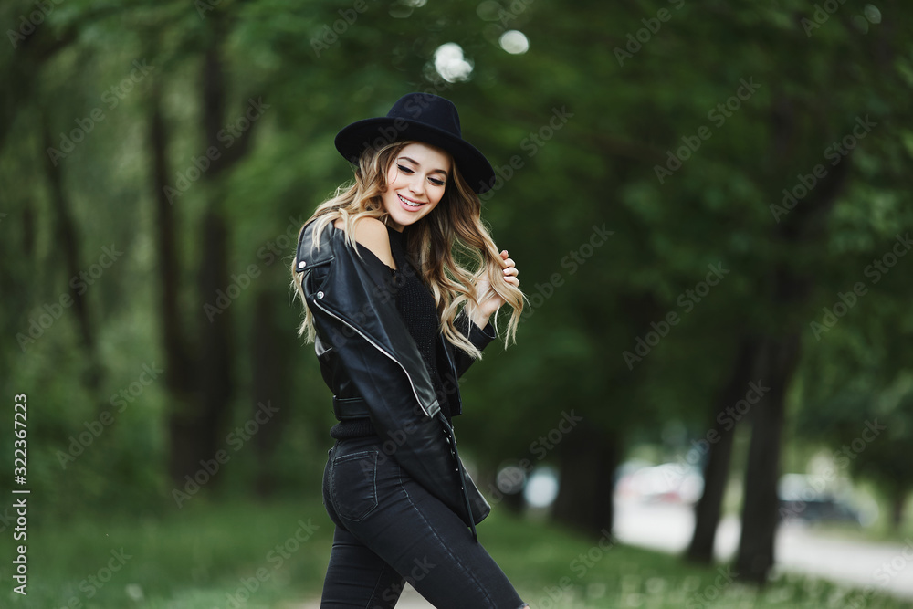 Happy blonde young woman with trendy makeup, in a black leather jacket and black hat, over a green background with bokeh effect and copy space by both sides. Fashion magazine concept. Fashion poster Happy blonde young woman with trendy makeup, in a black leather jacket and black hat, over a green background with bokeh effect and copy space by both sides. Fashion magazine concept. Fashion poster