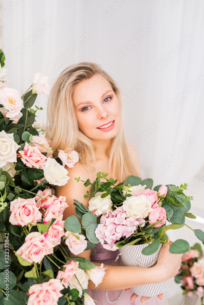 Beautiful romantic young woman with natural makeup posing against a background of flowers. Pale pink and white roses with greenery. A clean look and a snow-white smile. Perfume and cosmetic concept.