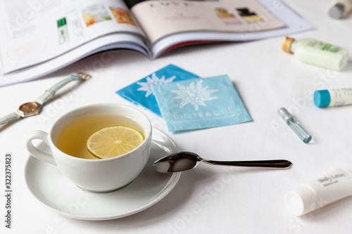White cup of green tea with lemon on a white bedsheet with aт illustrated magazine, tubes of cream, watch and tea bags. Healthy concept.