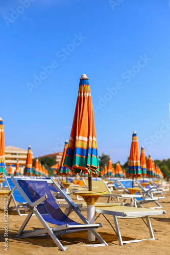 Blue sunbeds with bright multicolored orange green striped umbrellas on beautiful sand beach under blue sky. In front of adriatic sea. Summer holidays, vacation on the coast. Tourism in europe, Italy
