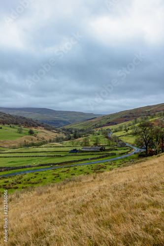 A scenic view of a mountain valley with grassy green slope, rueal road lane, trees and farm building under a stormy grey sky