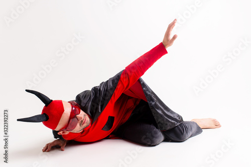 Fotografi Conceptual portrait of defeated and helpless devil man in black red vampire costume asking for help and mercy while lying on ground in studio and reaching his arm out