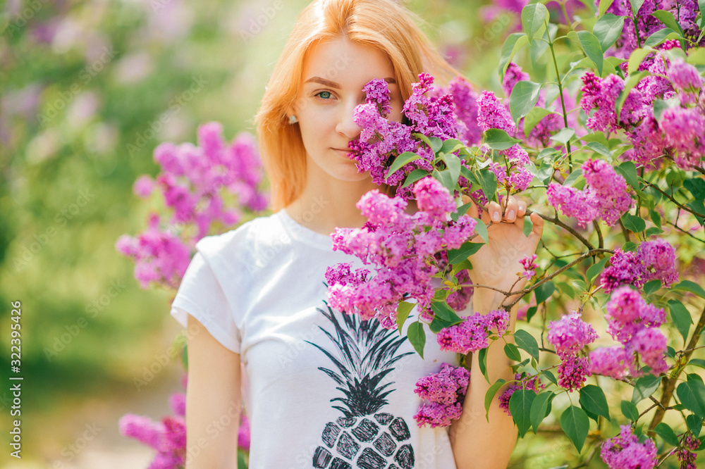 Fototapeta premium Handsome woman with red hair and pretty smile posing in lilac garden with flowers and enjoys the smell