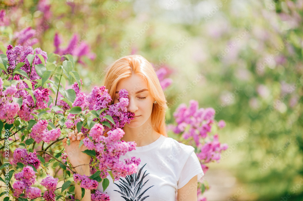 Fototapeta premium Pretty young female with red hair and fair skin smells lilac in the garden with closed eyes