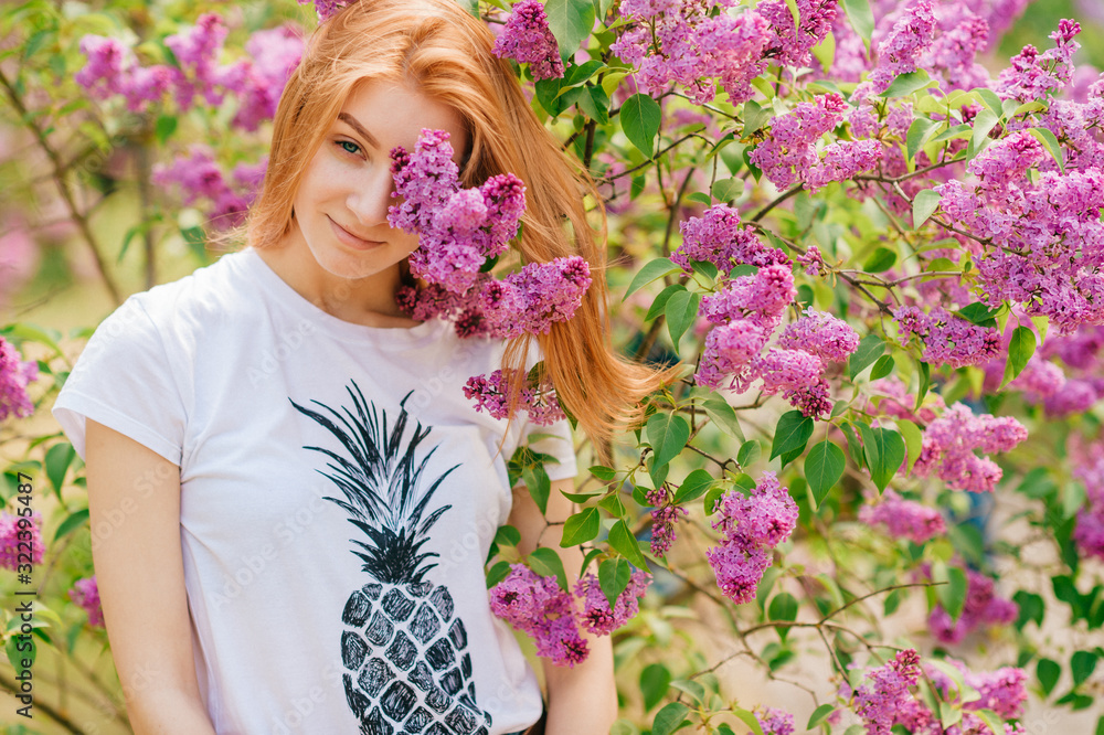 Handsome woman with red hair and pretty smile posing in lilac garden with flowers and enjoys the smell