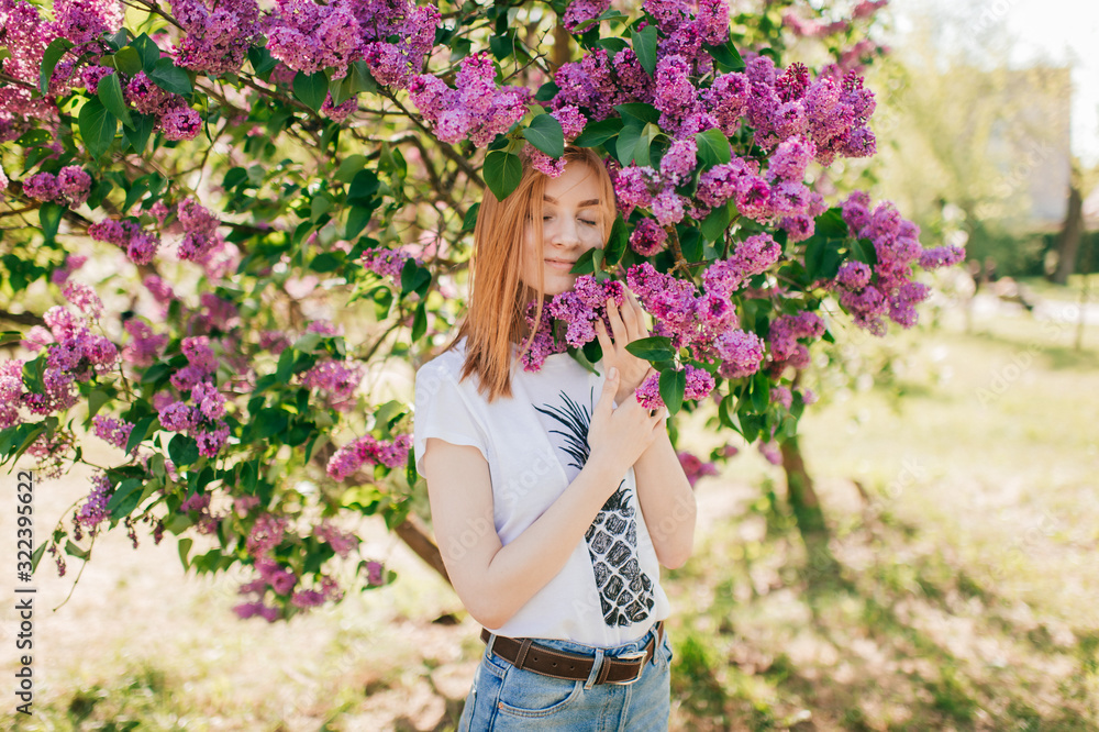 Pretty young female with red hair and fair skin smells lilac in the garden with closed eyes Pretty young female with red hair and fair skin smells lilac in the garden with closed eyes