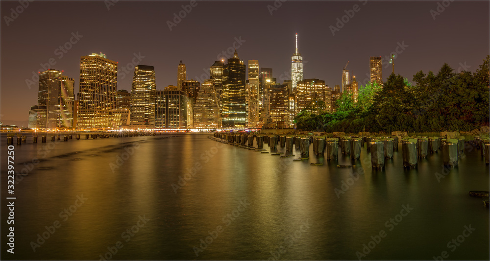 New York City skyline at night from Brooklyn Bridge Park with reflections in the smooth water.