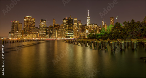 New York City skyline at night from Brooklyn Bridge Park with reflections in the smooth water.