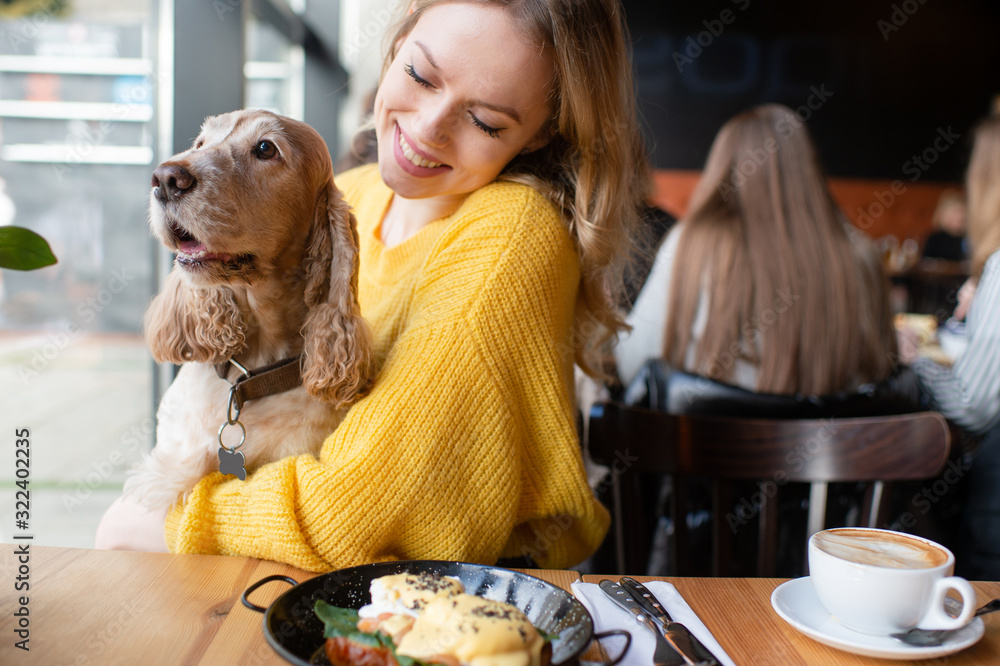 Φωτογραφία Young caucasian cheerful blonde girl sitting and hugging together with her lovely cocker spaniel dog in caffe at the table with lunch meal and cappuccino