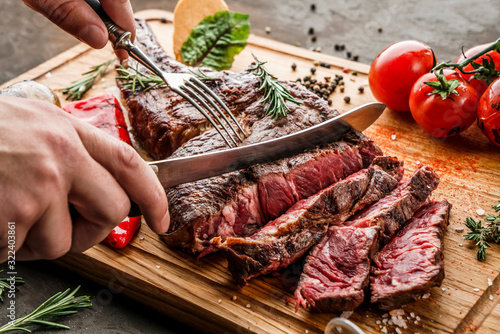 Hands cut grilled tomahawk meat medium rare or rib eye steak on wooden cutting board with grilled vegetables on dark background, close up