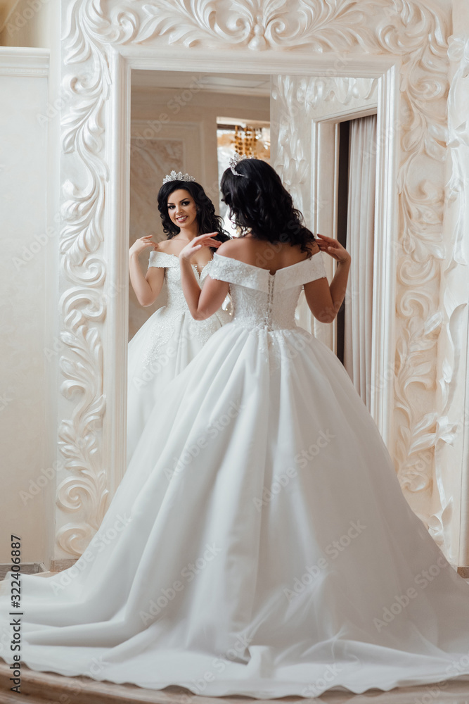 Amazing woman. Beautiful girl with long wavy hair. Brunette model with hairstyle. Attractive young bride in wedding dress in modern salon. Bride is looking into the mirror Amazing woman. Beautiful girl with long wavy hair. Brunette model with hairstyle. Attractive young bride in wedding dress in modern salon. Bride is looking into the mirror