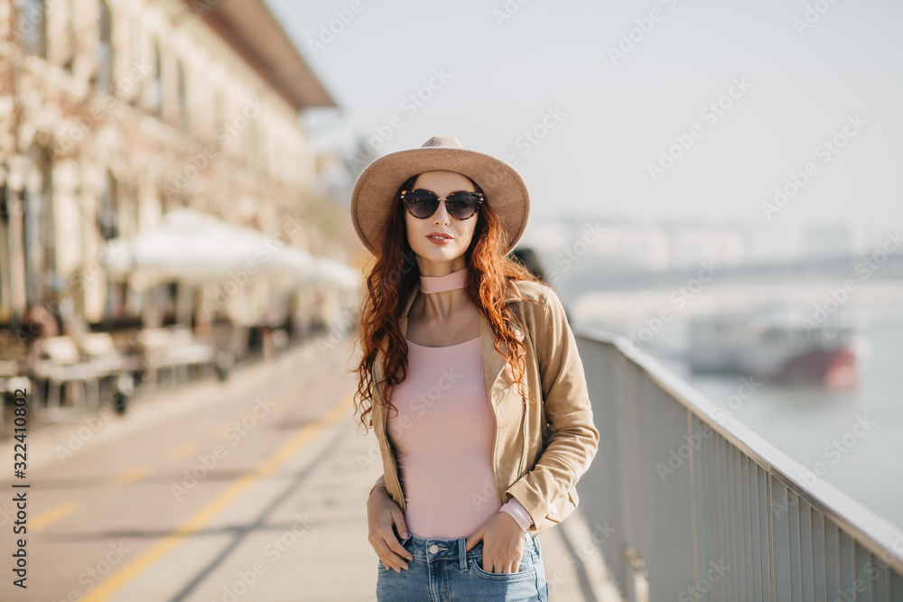 Gorgeous young woman in dark sunglasses standing in confident pose at embankment. Outdoor photo of glad ginger girl posing with hands in pockets.