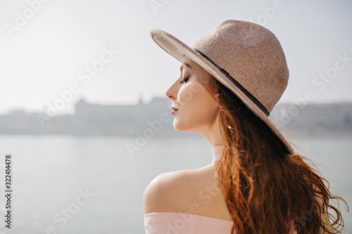 Close-up outdoor portrait of blissful ginger woman enjoying sea breeze. Photo of attractive female model with long dark hair in hat enjoying photoshoot on river. © Look!