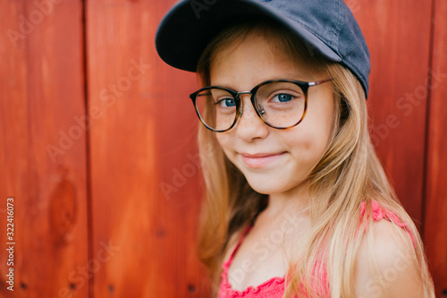 Little cute girl with blond hair and pink dress makes faces against the wooden wall