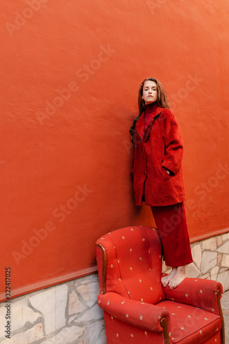 Portrait of young woman dressed in red standing barefoot on armrest of red lounge chair leaning on red wall photo