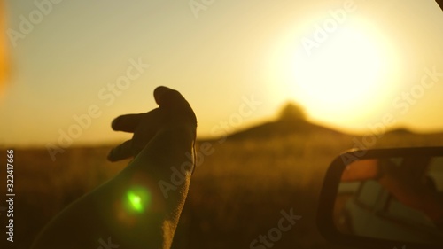 man traveler from the car window plays fingers with sunbeams. drivers hand is playing with sun from car window against beautiful sunset. Girl's hand waves sun. To travel by car