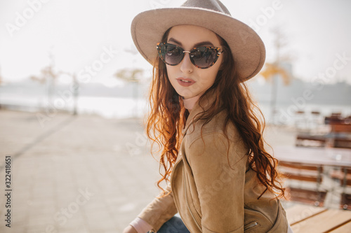 Magnificent ginger woman with serious face expression sitting on blur city background. Pretty girl in casual hat posing in good autumn day.