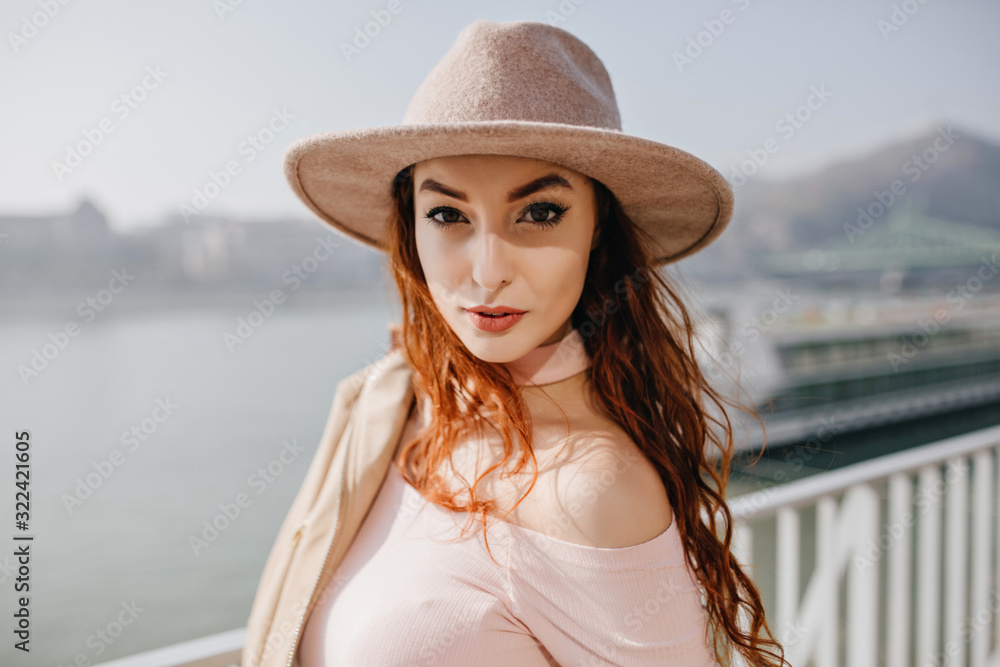 Gorgeous long-haired ginger girl with serious face expression standing on ocean background. Outdoor photo of carefree european lady in beige fedora chilling near sea.