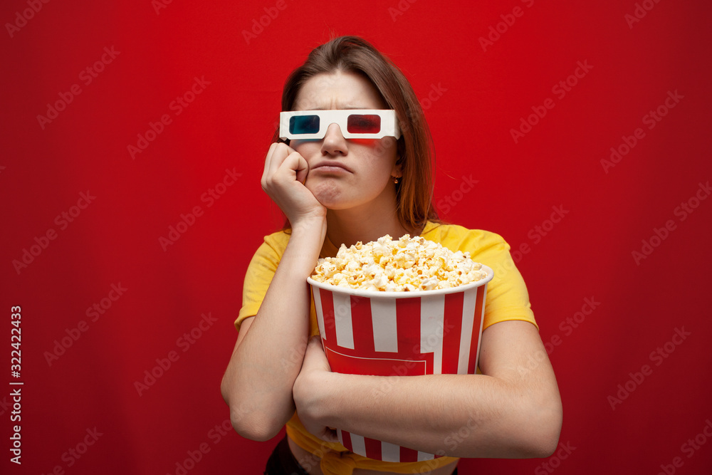 sad young girl watching a bad, boring movie in 3d glasses and with popcorn on a red background