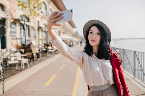 Happy dark-haired girl in romantic outfit making selfie near street cafe. Outdoor photo of refined caucasian female model in hat taking picture of herself.