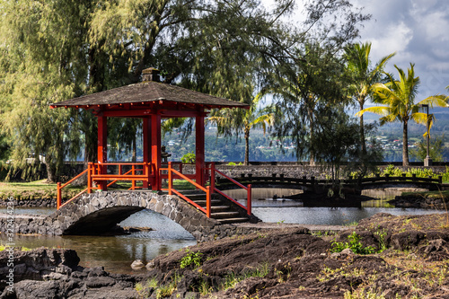 Hilo, Hawaii, USA. - January 9, 2012: Closeup of Japanese black and red small bow bridge over pond in Liliuokalani Gardens. Green trees and black rocks under blue sky with big white clouds.
