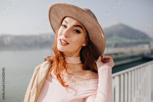 Stunning white woman in fedora relaxing near sea in cold day. Outdoor portrait of ginger refined girl touching hat on river background.