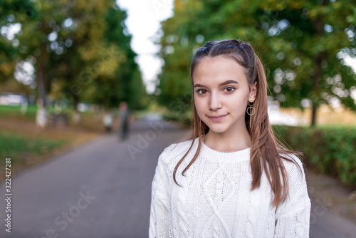 Portrait of a beautiful schoolgirl girl, summer park, free space for copy text, casual clothes, white sweater, long hair, positive emotions, smiling and resting after school.