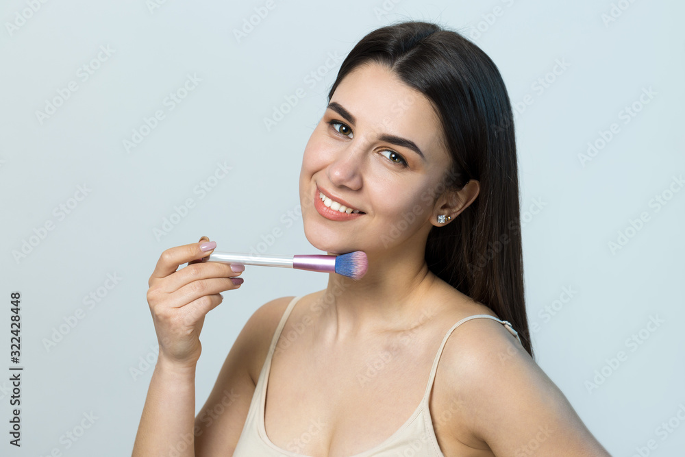 Close-up of a young girl in a light top on a white background making a facial make-up. A pretty woman holds a cosmetic brush near her face and smiles. Close-up of a young girl in a light top on a white background making a facial make-up. A pretty woman holds a cosmetic brush near her face and smiles.