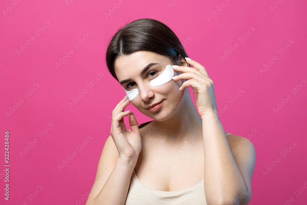 Close up portrait of pretty attractive girl with naked shoulders using patches under eyes. Standing over pink background. Close up portrait of pretty attractive girl with naked shoulders using patches under eyes. Standing over pink background.