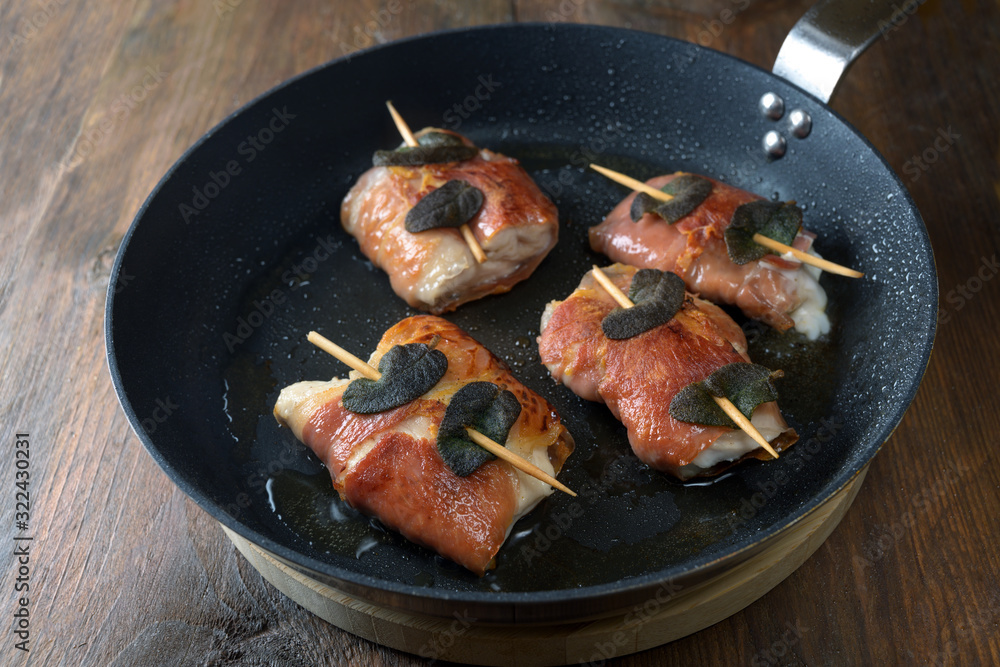 Roasted chicken saltimbocca wrapped with air-dried ham and sage leaves in a black pan on a rustic wooden table Roasted chicken saltimbocca wrapped with air-dried ham and sage leaves in a black pan on a rustic wooden table