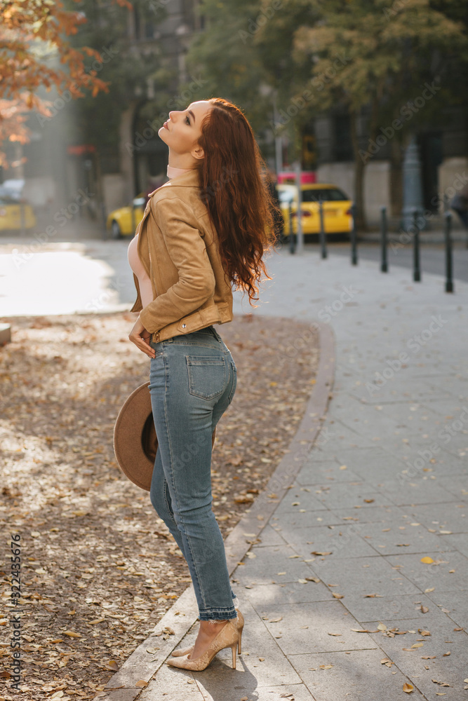 Shapely ginger girl in casual jeans looking up during photoshoot. Outdoor shot of slim red-haired lady enjoying autumn day. Shapely ginger girl in casual jeans looking up during photoshoot. Outdoor shot of slim red-haired lady enjoying autumn day.
