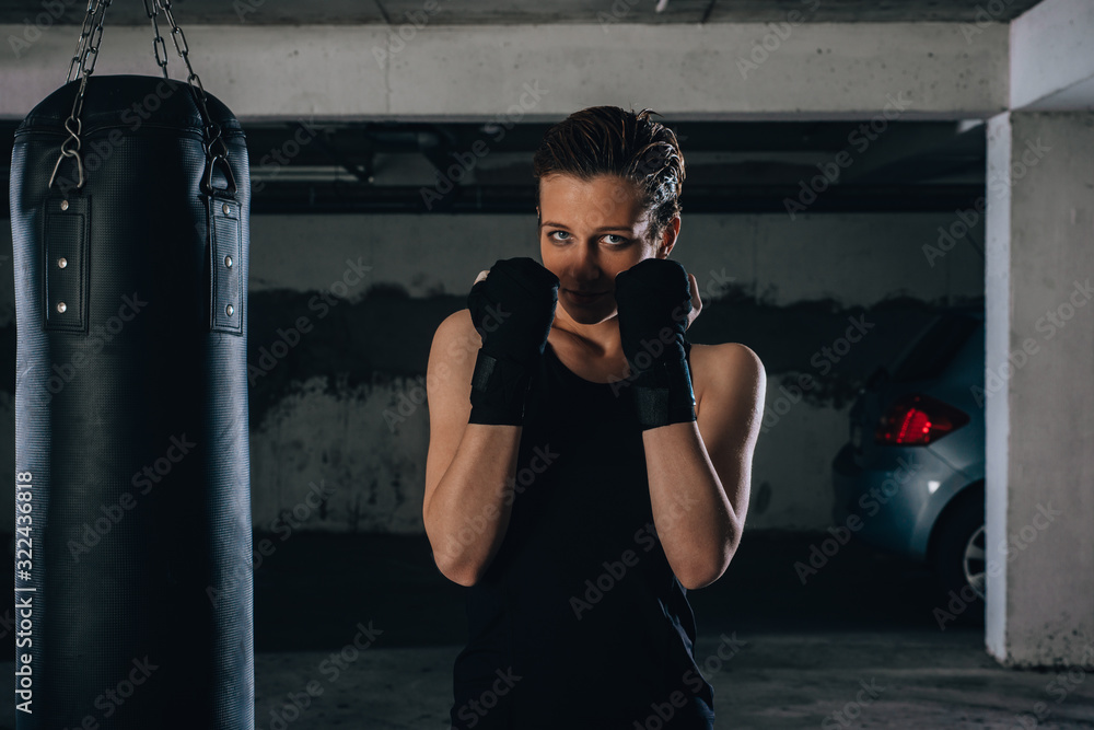 Portrait of a young strong woman winding black bandages on the wrists, preparing for boxing in a garage.