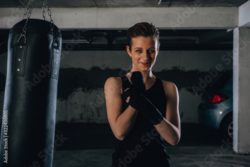 Portrait of a young strong woman winding black bandages on the wrists, preparing for boxing in a garage.