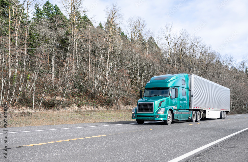 Big rig modern green bonnet semi truck with refrigerator semi trailer running on the road with forest on the hillside Big rig modern green bonnet semi truck with refrigerator semi trailer running on the road with forest on the hillside