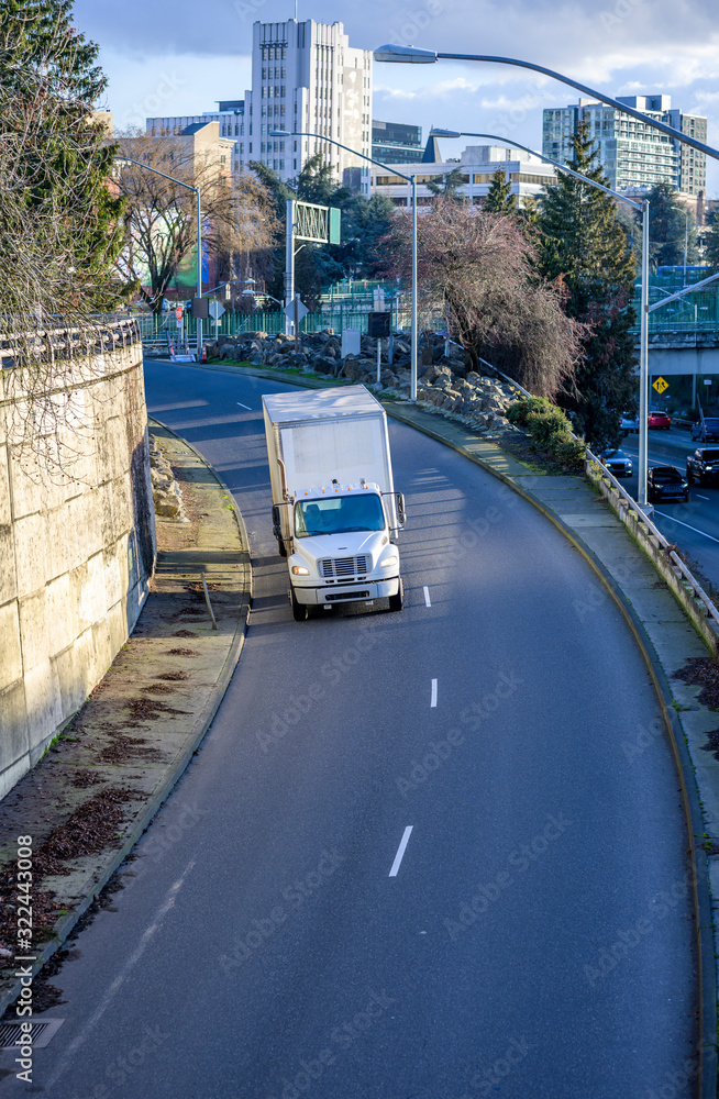 Obraz premium Medium duty rig white semi truck transporting goods in box trailer going to highway entrance running from the city street downhill