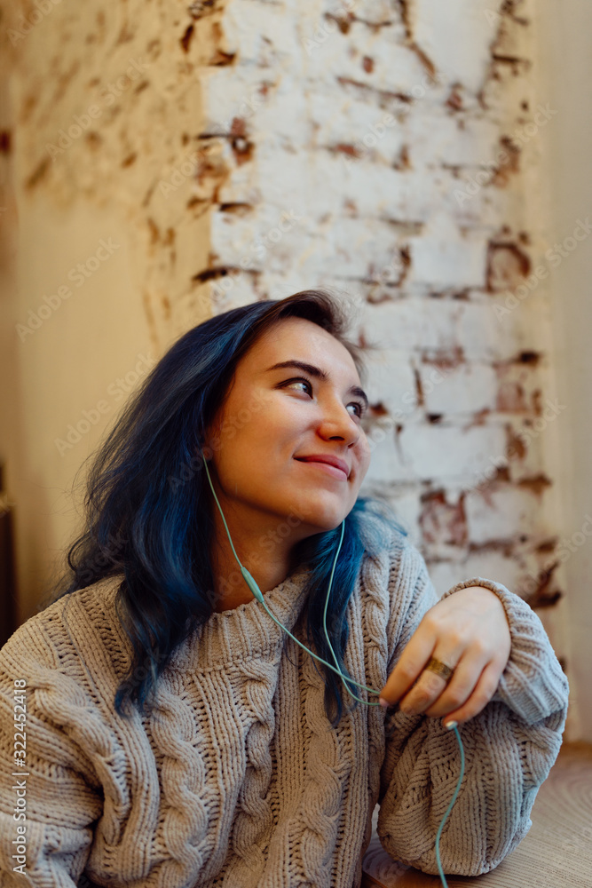 Fototapeta premium Young adult female with dyed blue hair listening music in a coffee shop during blue hour, shallow selective focus