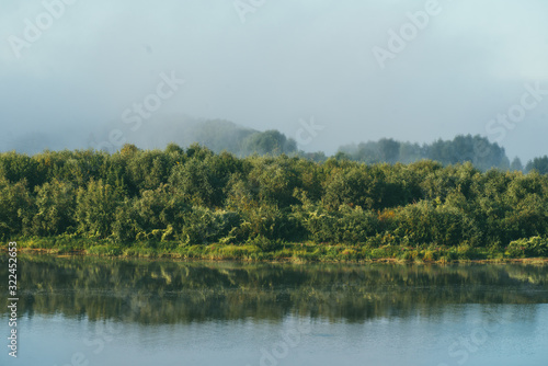 Early morning l Spring landscape with a full flowing river in the foreground and a green Bank overgrown with bushes and trees in the background. The distant plan is drowned in a thick morning fog
