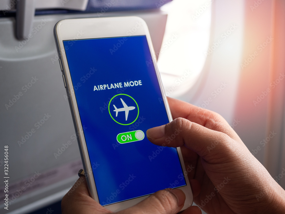 Flight mode concept. Hand holding white smartphone and turned on airplane mode on screen near the window on the airplane.