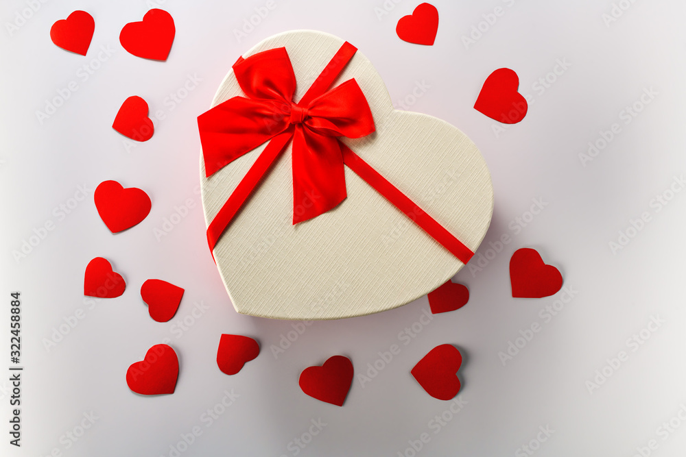 Box. Heart-shaped gift box with red ribbon stands in the middle on a white background with little red cardboard hearts. The view from the top. Valentine's day and Birthday Box. Heart-shaped gift box with red ribbon stands in the middle on a white background with little red cardboard hearts. The view from the top. Valentine's day and Birthday