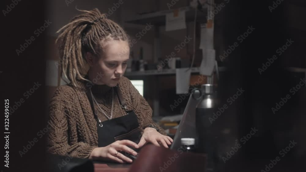 Young stylish woman with dreadlocks, in a voluminous sweater, sits in front of a laptop in a dark room or workshop. She is looking for examples for inspiration or communicating with customers. Young stylish woman with dreadlocks, in a voluminous sweater, sits in front of a laptop in a dark room or workshop. She is looking for examples for inspiration or communicating with customers.