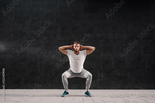 Full length of attractive caucasian dedicated bearded man in tracksuit doing squats outside. In background is gray wall.