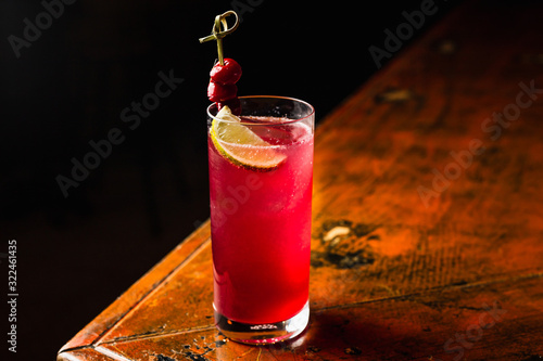 A red cocktail in a highball glass garnished with lemon and cherries