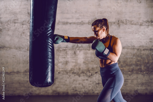 Full length of young dedicated caucasian muscular female boxer in sportswear with ponytail and boxing gloves punching boxing bag while standing in the gym.