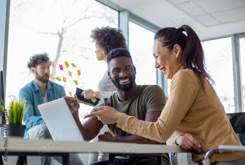 Photos A man and a woman laughing while looking at the lap top with another man and a woman in the background of a workspace environment