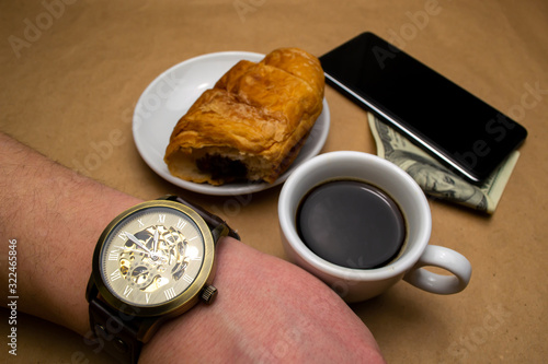 Wristwatch on hand white cup with coffee croissant mobile phone dollar on a beige background