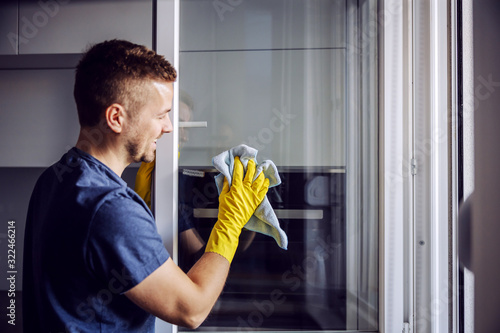 Young bearded smiling positive man with rubber gloves cleaning window with cloth. After rain there is always stains on glass. photo